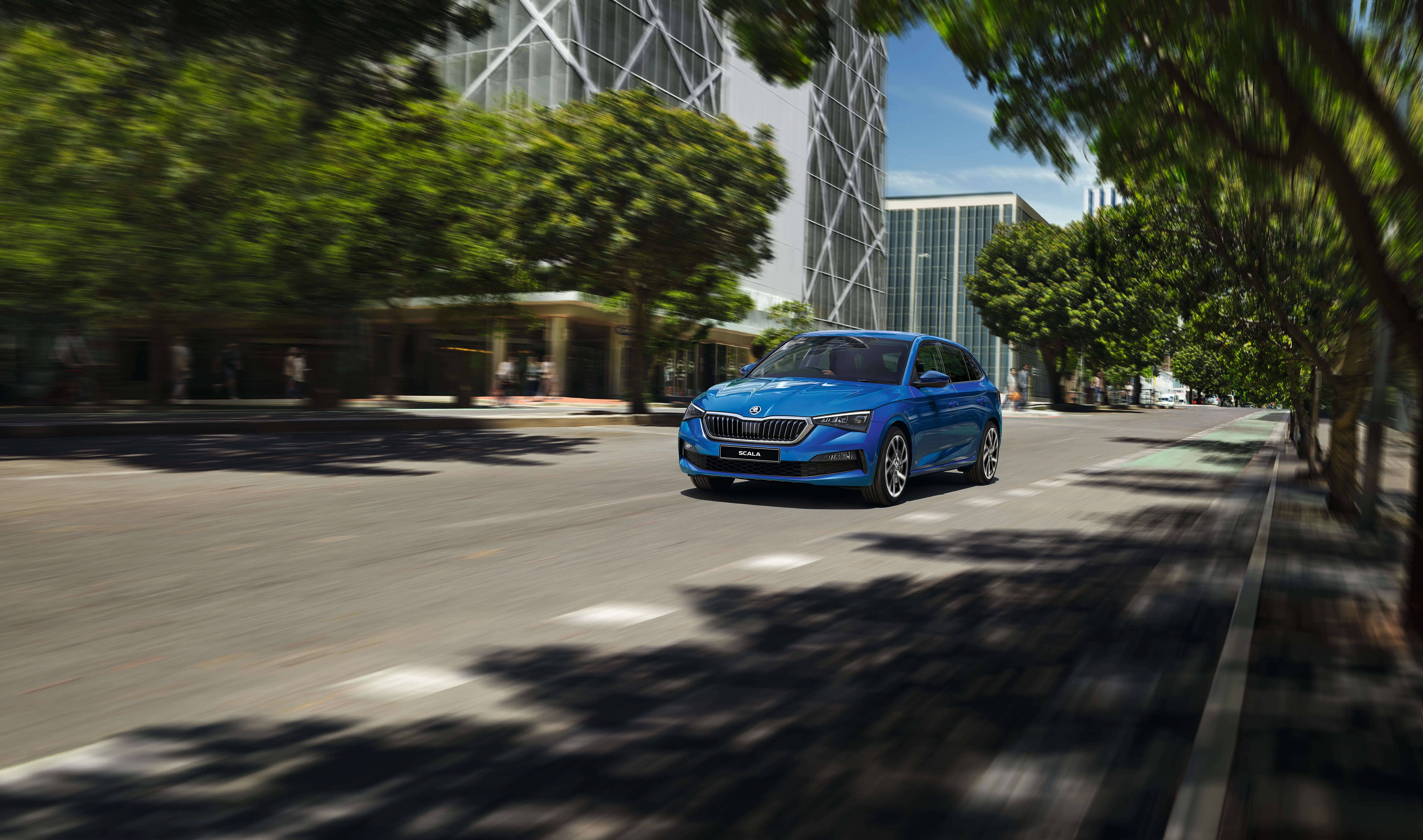 Blue Škoda Scala driving through a modern city street, showcasing its sleek design, LED headlights, and urban-friendly performance in a vibrant urban setting.