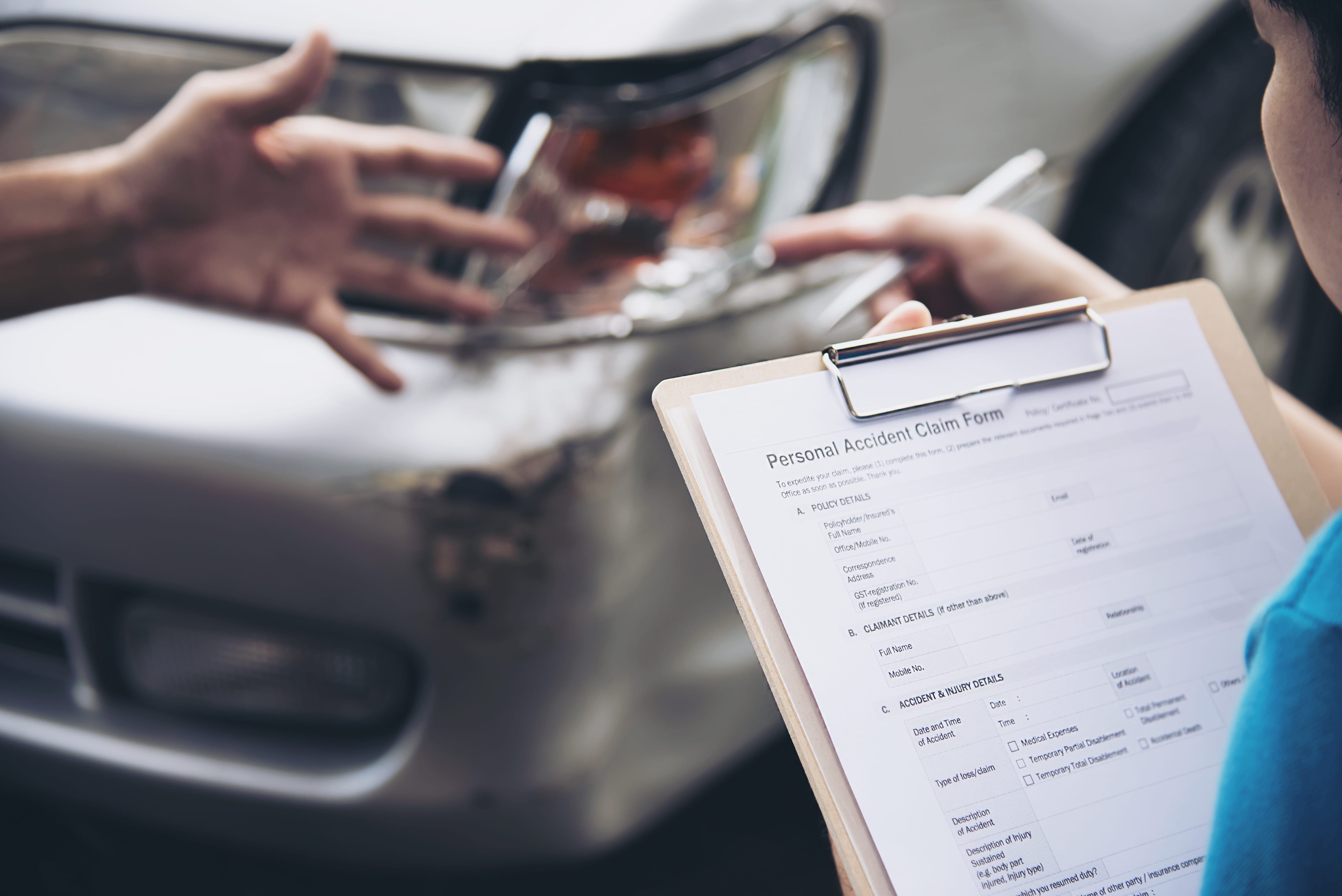 Insurance agent holding a clipboard with a personal accident claim form while inspecting a damaged car front bumper after a collision, highlighting car insurance costs and the importance of safety features.